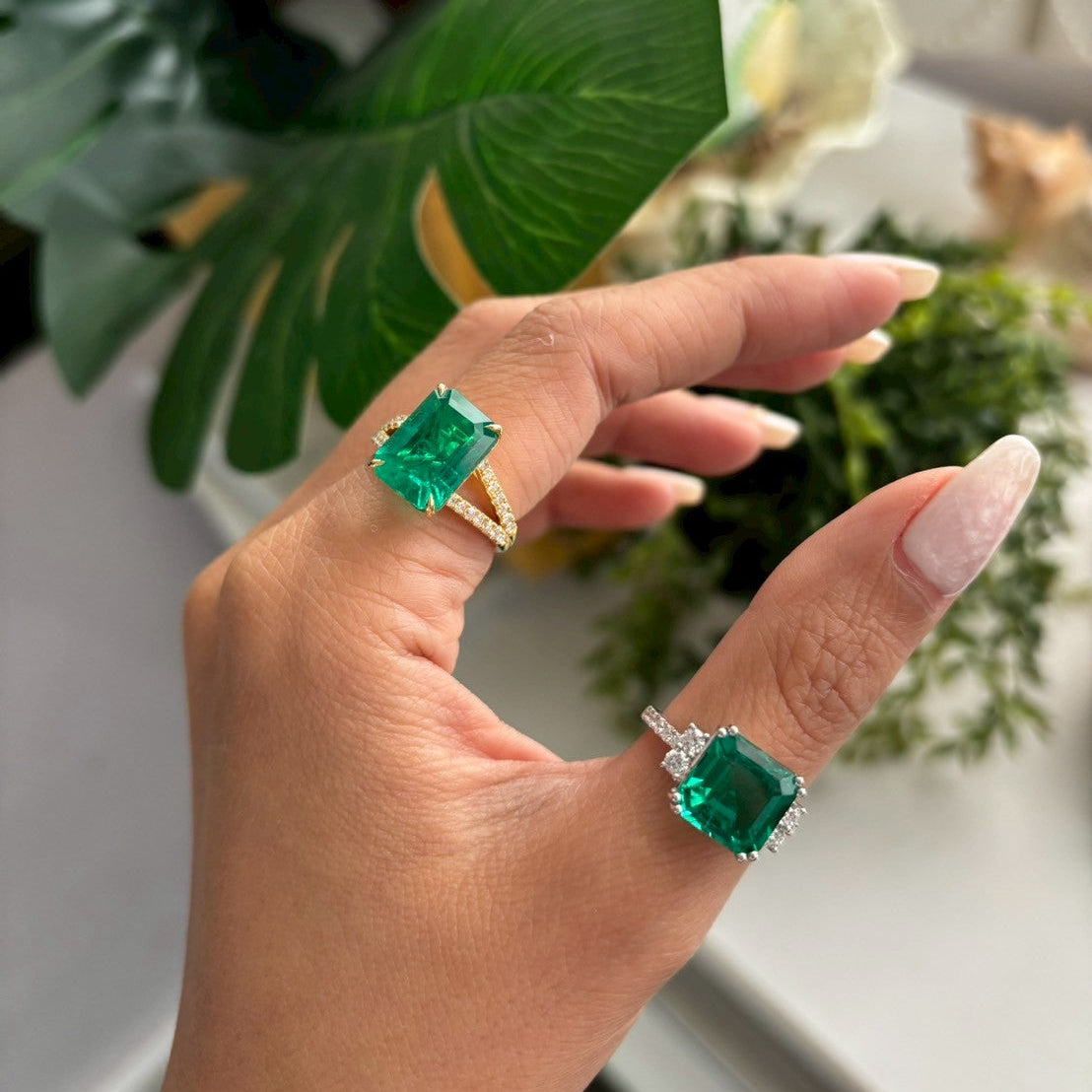 Hand wearing two emerald green rings with diamond accents against a blurred natural background